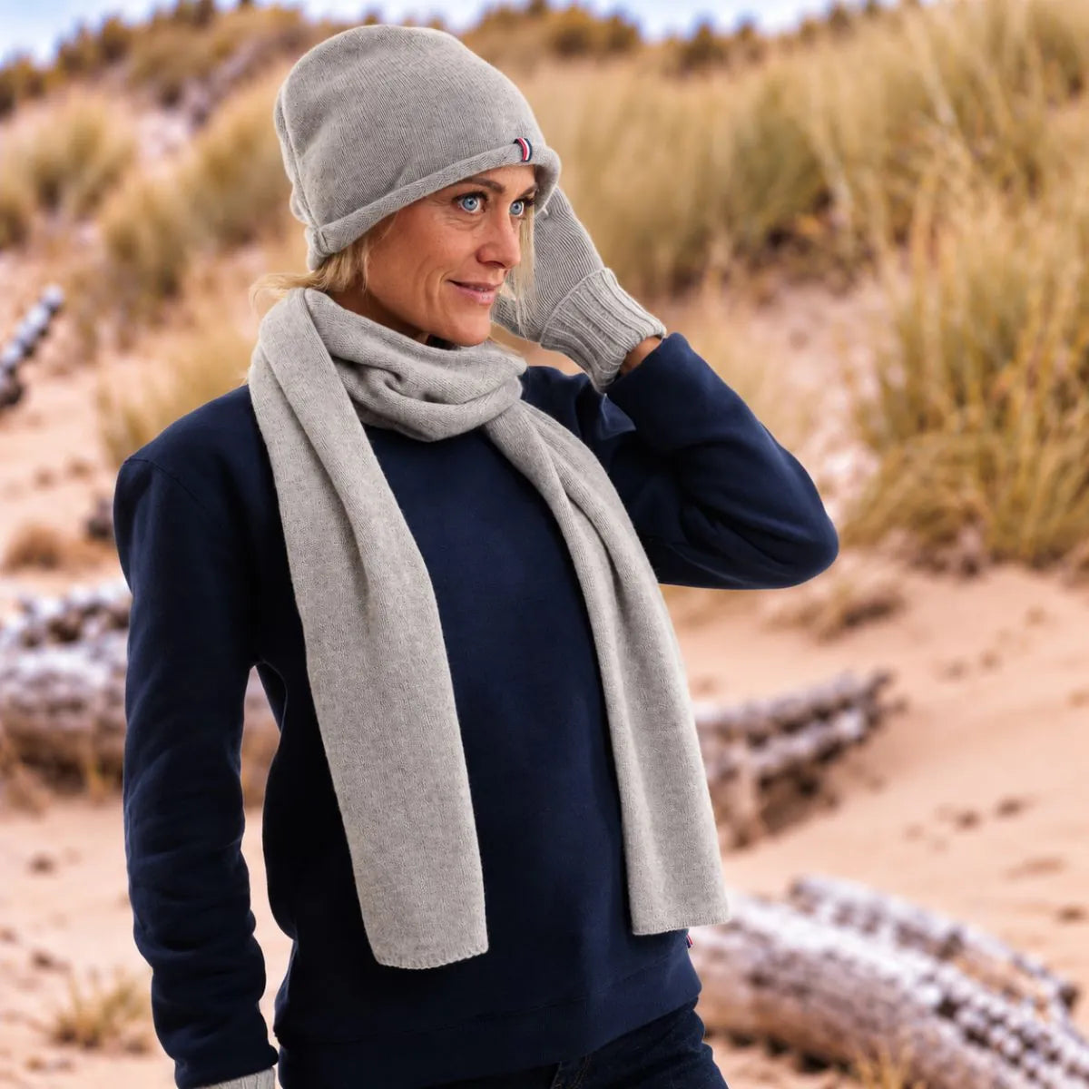 Femme blonde dans les dunes portant le bonnet tombant en cachemire gris, l'écharpe et les gants assortis