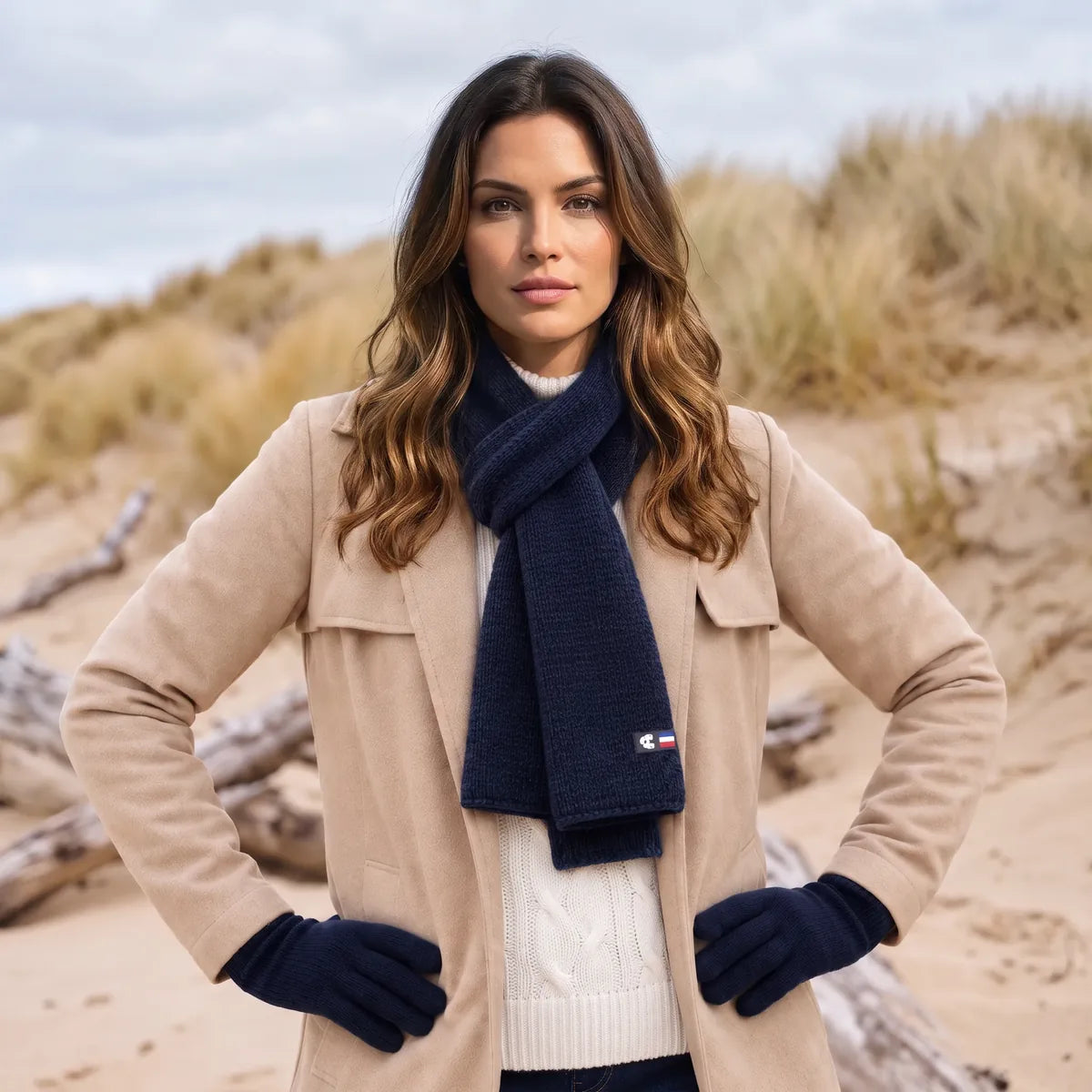 Femme en manteau beige portant l'écharpe en cachemire bleu marine nouée et les gants assortis, dans les dunes