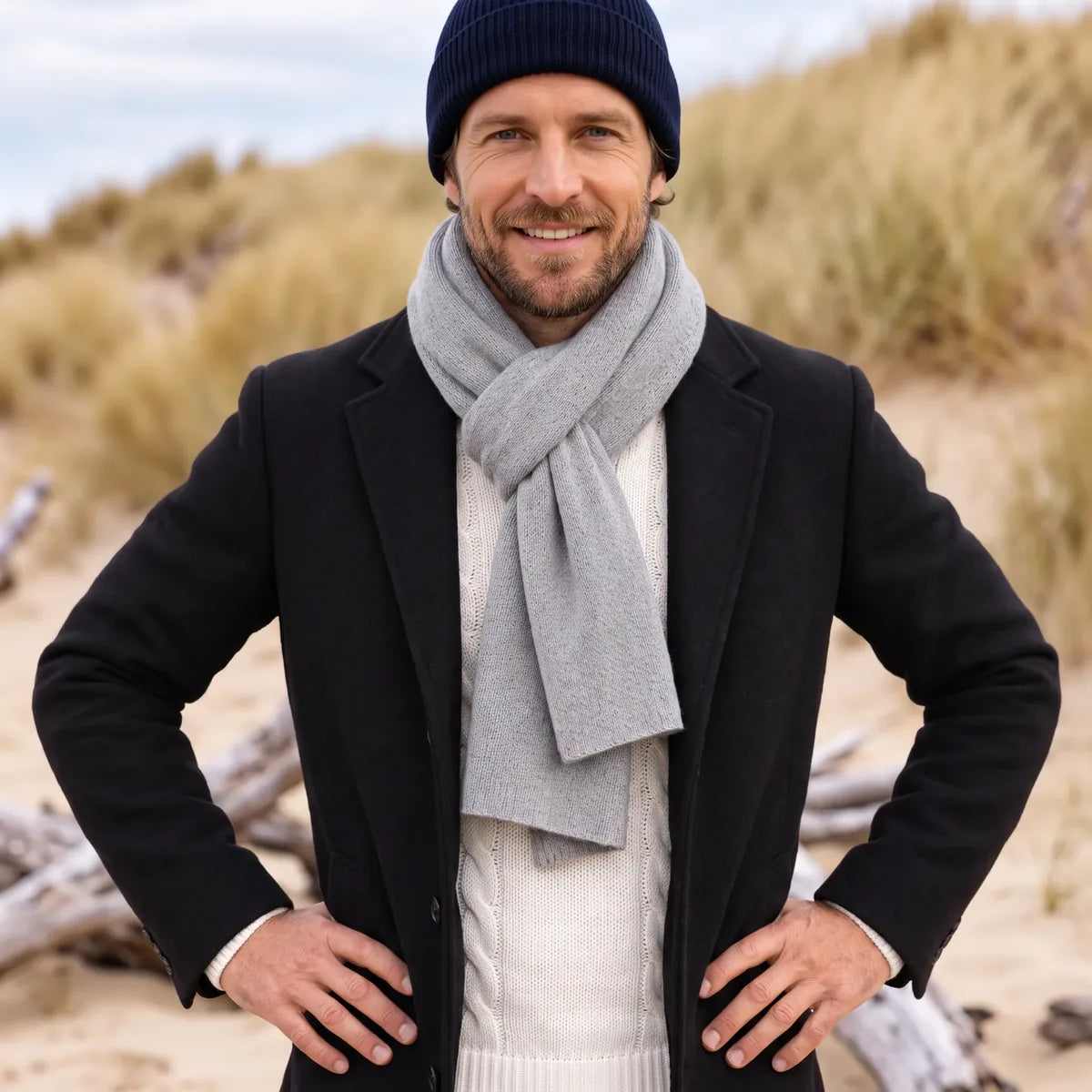 Homme souriant portant l'écharpe en cachemire grise nouée sur un manteau noir et un pull écru, dans les dunes