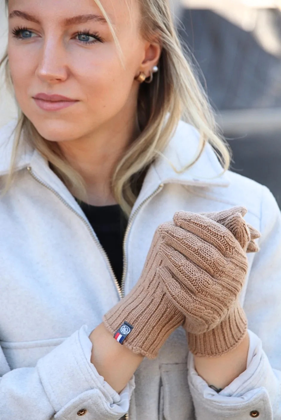 Femme portant des gants torsadés camel en laine mérinos, mains jointes devant un manteau clair
