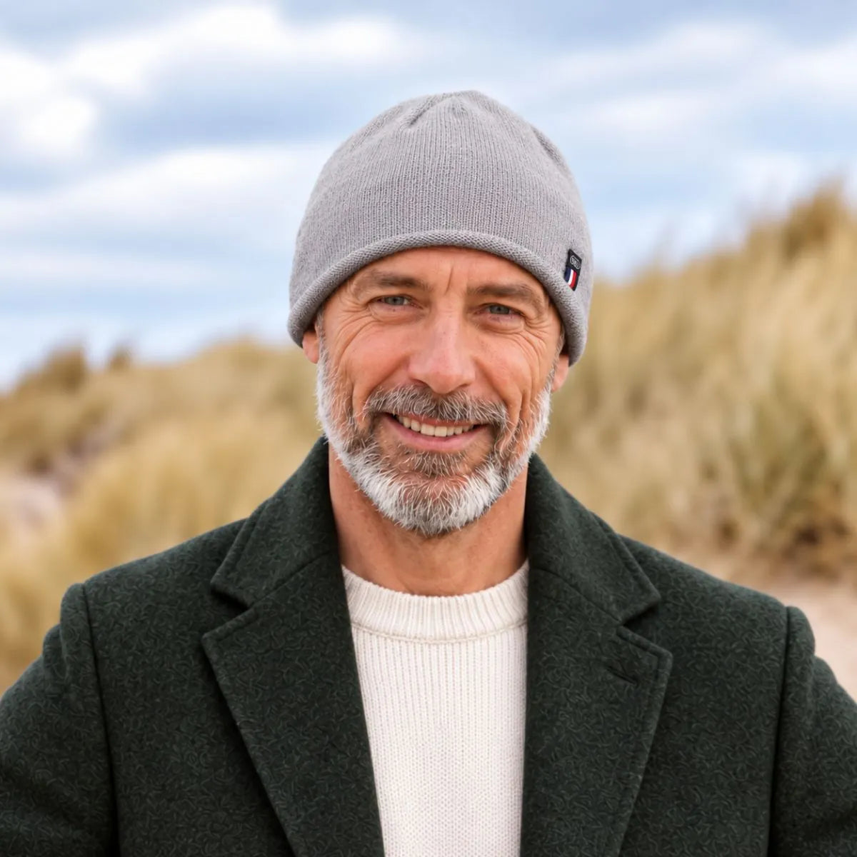 Homme barbu en manteau vert foncé portant le bonnet en cachemire gris, dans les dunes