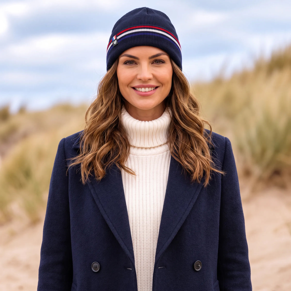 Femme en manteau bleu marine et col roulé crème portant le bonnet bleu marine à revers tricolore, dans les dunes
