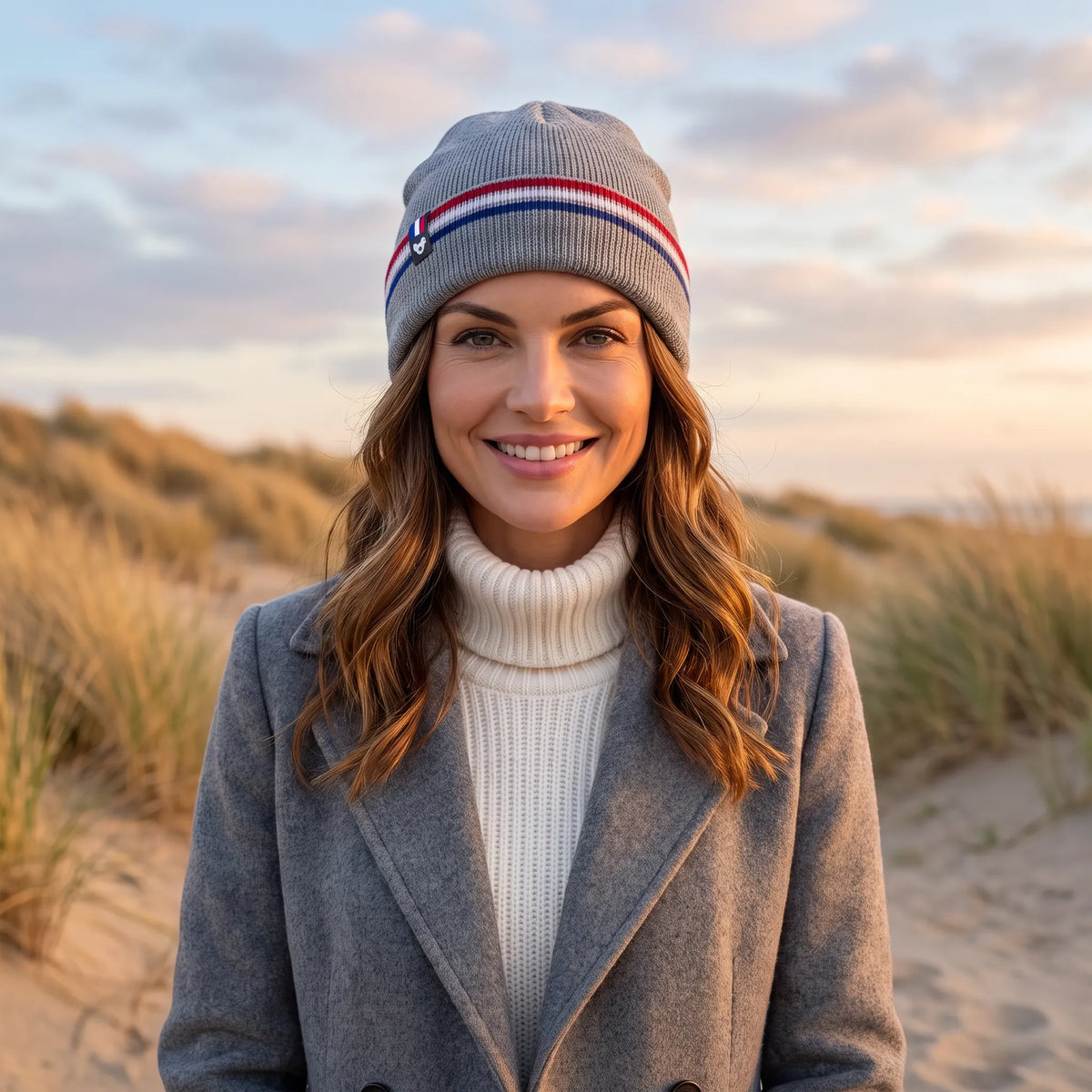 Femme souriante en manteau gris et col roulé crème portant le bonnet gris à revers tricolore, dans les dunes