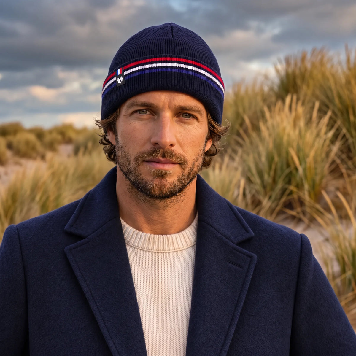 Homme en manteau bleu marine et pull écru portant le bonnet bleu marine à revers tricolore, dans les dunes