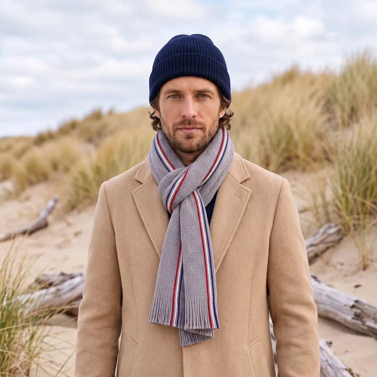 Écharpe tricolore grise nouée autour du cou d'un homme en manteau camel et bonnet bleu marine, devant des dunes